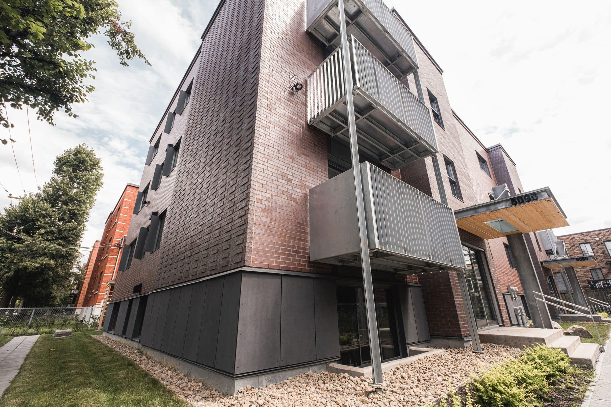 Coin de l’immeuble : brique, panneaux métalliques foncés et balcons